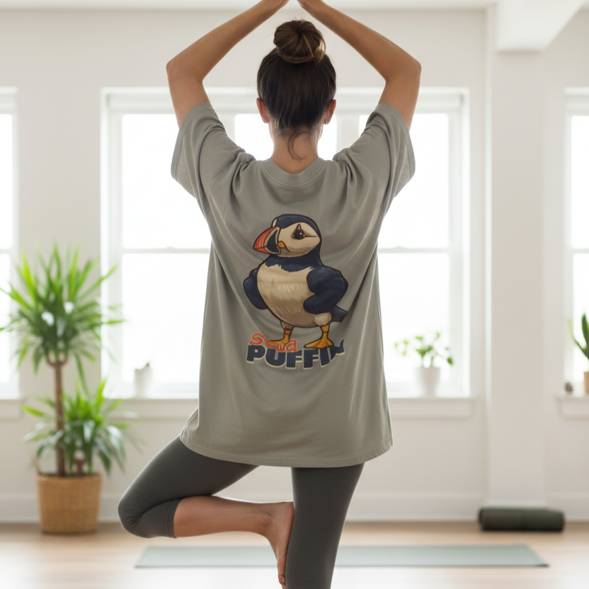 A woman practices yoga in a sunlit room, standing in tree pose. She wears a gray back print shirt with a cartoon puffin on the back. The mood is calm and focused.