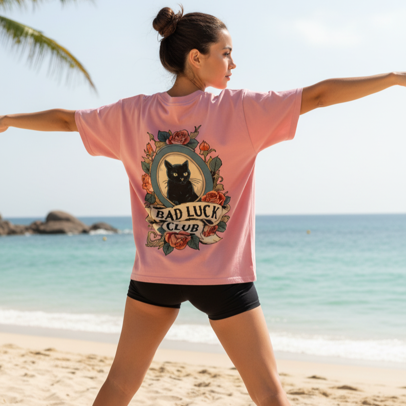 Woman wearing a pink backprint tattoo t-shirt with a black cat graphic and 'Bad Luck Club' text on a beach.