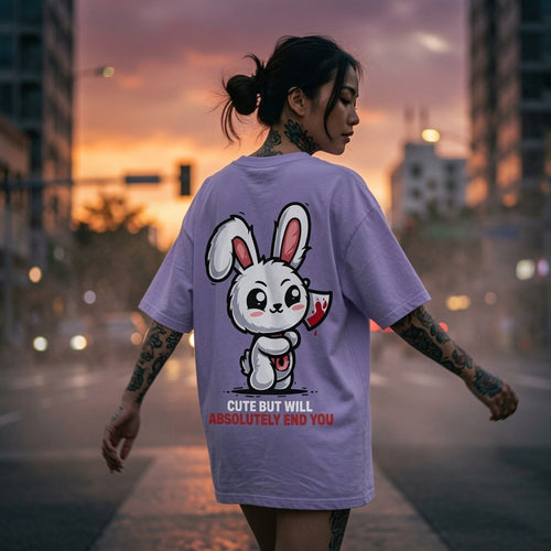 Person with tattoos wearing a lavender T-shirt featuring a cute cartoon bunny holding a bloody knife, with text: "Cute but will absolutely end you." The setting has an urban street at sunset, creating a dramatic contrast between the playful graphic and the edgy message.