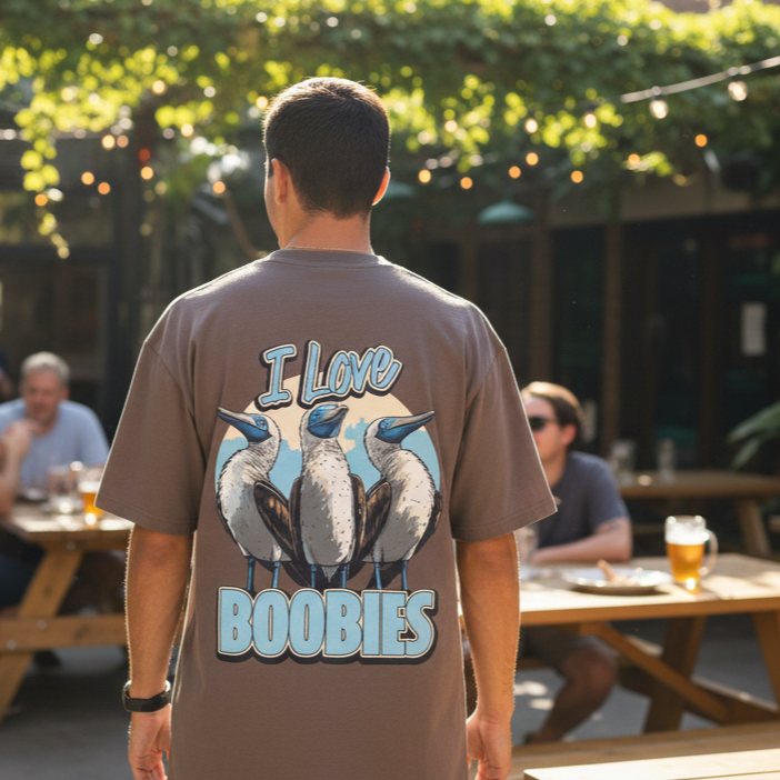 A man stands outdoors in a pub beer garden wearing a graphic back print t-shirt with an illustration of blue-footed boobies and the words "I Love Boobies" on the back. People sit at picnic tables in the background.